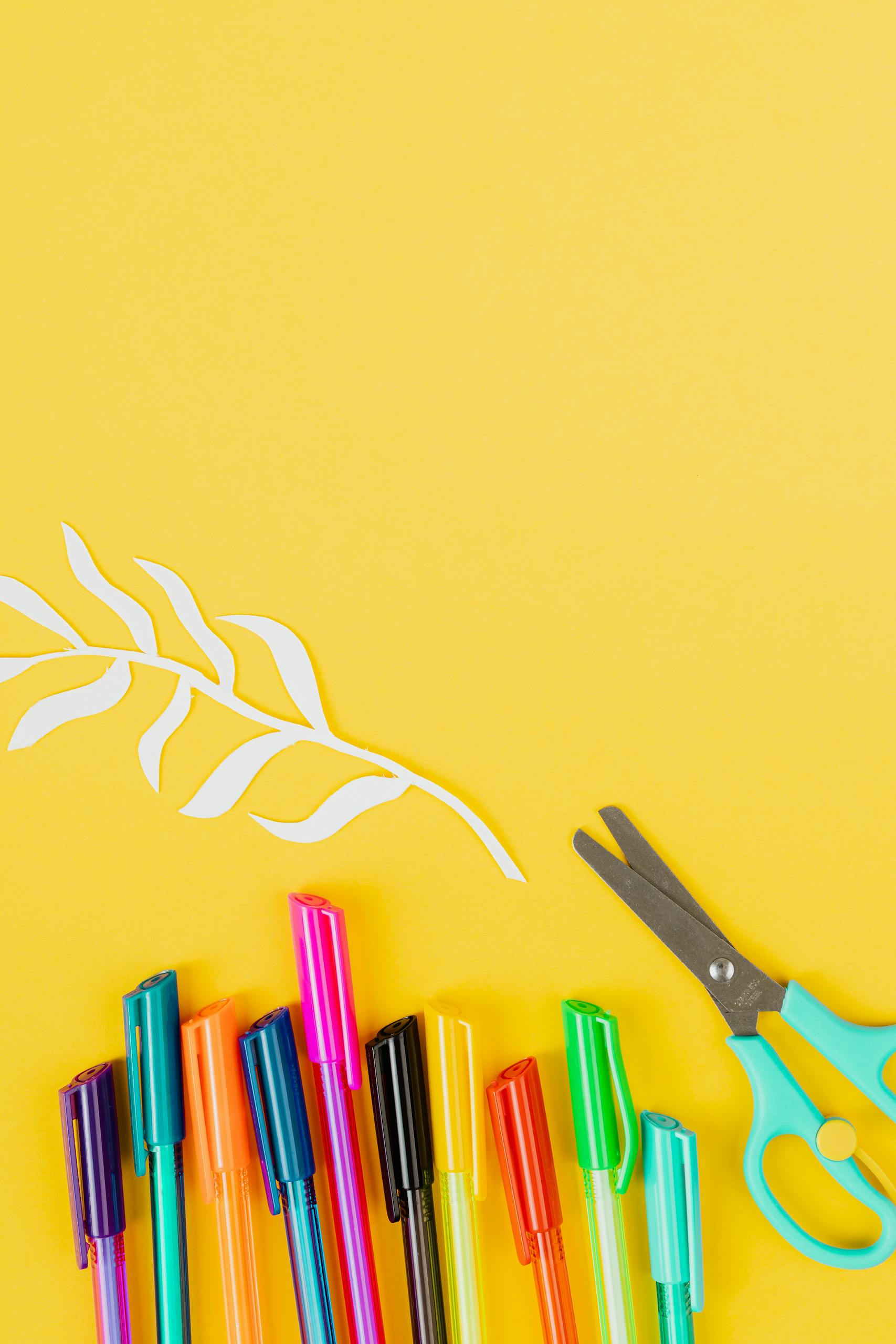 A vibrant arrangement of colorful pens and scissors against a yellow background with paper cutouts.