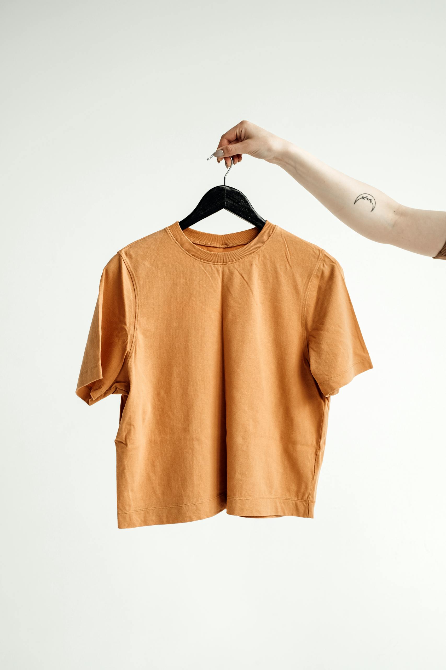 Minimalist shot of an orange t-shirt on a hanger held by a tattooed hand against a white background.